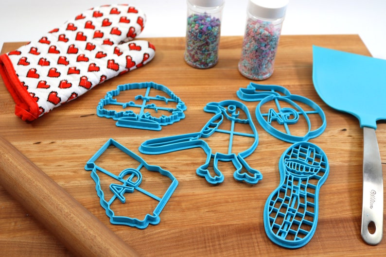 Puede incluir: Un juego de cortadores de galletas de pl&aacute;stico azul en forma de &aacute;rbol, p&aacute;jaro, cacahuete, contorno de estado con las letras "AD" dentro y un c&iacute;rculo con las letras "AD" dentro. Tambi&eacute;n hay un rodillo, una esp&aacute;tula y dos frascos de chispas.