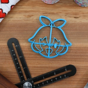 May include: A blue plastic cookie cutter shaped like a cartoon girl with a bow in her hair. The girl is winking and smiling.