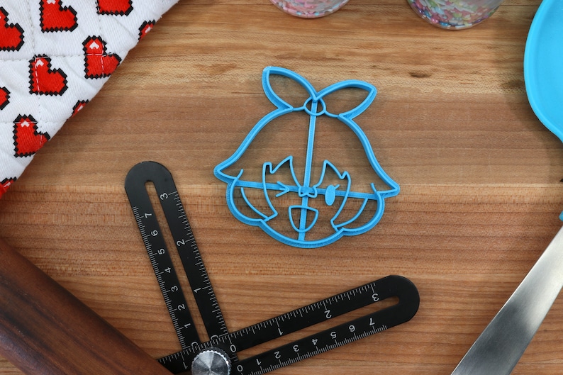 May include: A blue plastic cookie cutter shaped like a cartoon girl with a bow in her hair. The girl is winking and smiling.