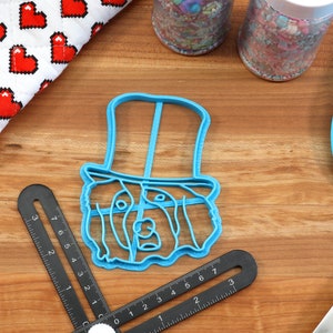 May include: A blue plastic cookie cutter in the shape of a dog wearing a top hat sits on a wooden surface. Other items include a red and white heart-patterned oven mitt, sprinkles, and a blue spatula. A black angle finder is also present.