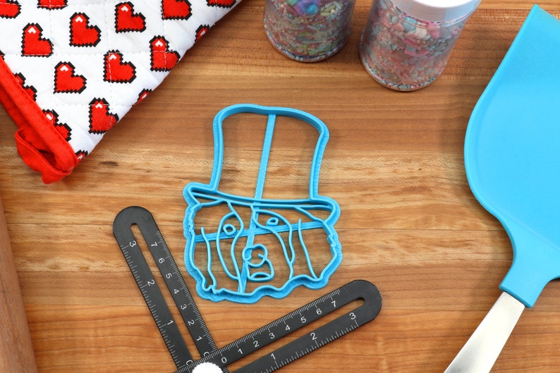 May include: A blue plastic cookie cutter in the shape of a dog wearing a top hat sits on a wooden surface. Other items include a red and white heart-patterned oven mitt, sprinkles, and a blue spatula. A black angle finder is also present.