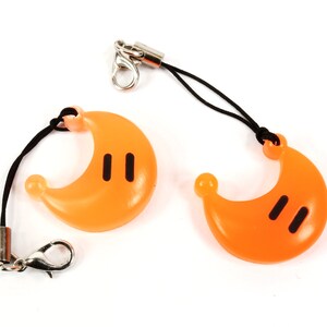May include: Two orange crescent moon shaped charms with black eyes on a black string with a silver clip.