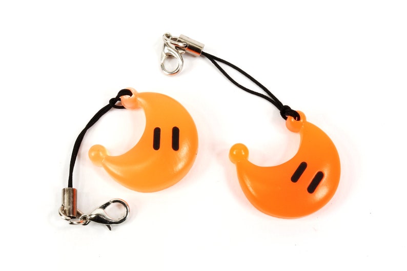 May include: Two orange crescent moon shaped charms with black eyes on a black string with a silver clip.