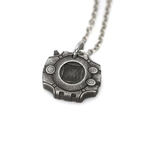Puede incluir: Un collar de plata con un colgante con forma de dispositivo circular con una ventana cuadrada en el centro. El colgante tiene una superficie texturizada y un dise&ntilde;o detallado.