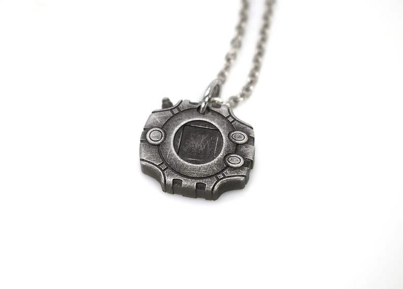 Puede incluir: Un collar de plata con un colgante con forma de dispositivo circular con una ventana cuadrada en el centro. El colgante tiene una superficie texturizada y un dise&ntilde;o detallado.