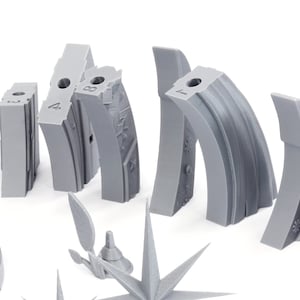 May include: An assortment of gray 3D-printed architectural model pieces, including curved and angular forms, with numbers and decorative details. The pieces are arranged against a white background, suggesting a design or construction theme.