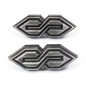 May include: Two metal geometric shaped pins with a brushed silver finish. Each pin has a repeating pattern of lines and angles, creating an abstract design. The pins are displayed on a white background.