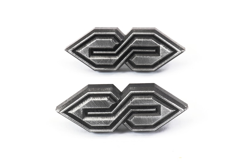 May include: Two metal geometric shaped pins with a brushed silver finish. Each pin has a repeating pattern of lines and angles, creating an abstract design. The pins are displayed on a white background.