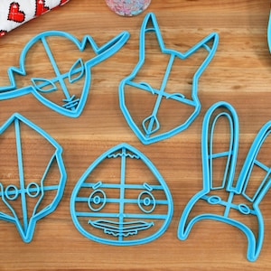 May include: Five blue plastic cookie cutters shaped like cartoon characters. The characters include a fox, a rabbit, an alien, a bird, and a creature with large eyes.