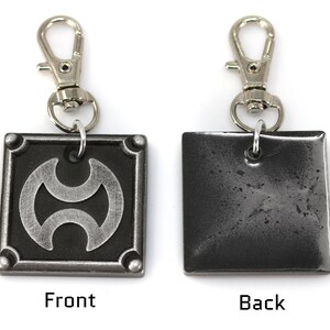 May include: A silver metal keychain with a square charm featuring a stylized symbol on the front and a plain back. The keychain has a silver spring clasp.