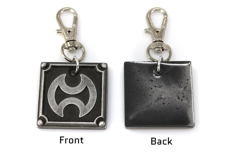 May include: A silver metal keychain with a square charm featuring a stylized symbol on the front and a plain back. The keychain has a silver spring clasp.