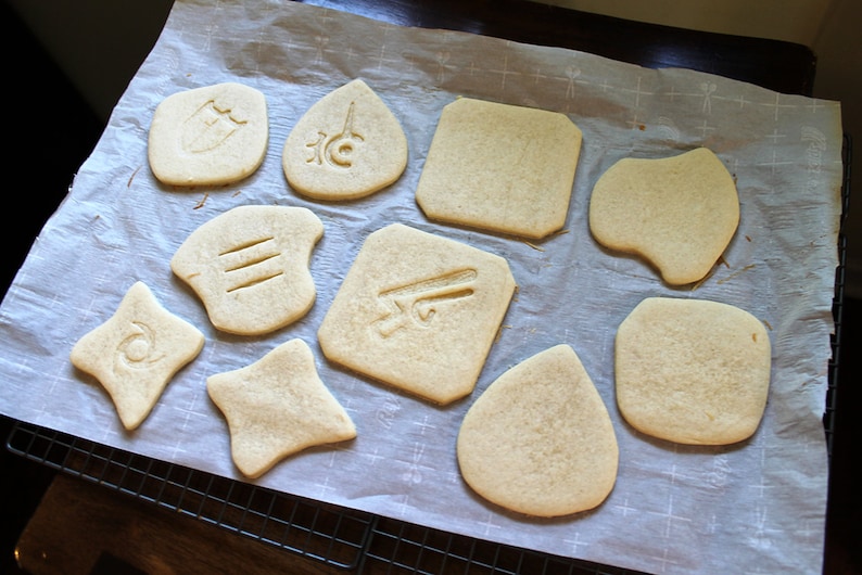 Op de afbeelding: Een vel bakpapier met negen eenvoudige suikerkoekjes, elk met een ander ontwerp dat eruit is gesneden. De ontwerpen omvatten een schild, een traan, een vierkant, een hart, een rechthoek, een traan, een ster, een ruit en een rechthoek.