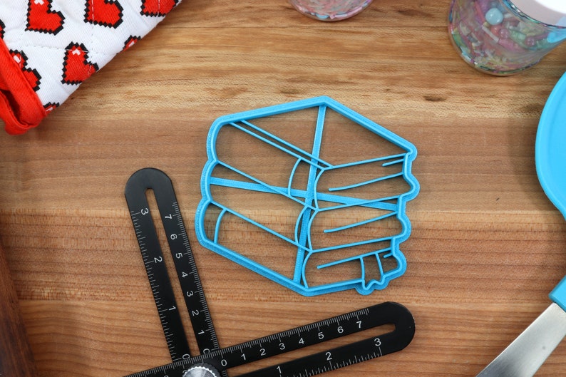 K&ouml;nnte beinhalten: Ein blauer Plastik-Ausstecher in Form eines B&uuml;cherstapels. Der Ausstecher liegt auf einer Holzoberfl&auml;che.