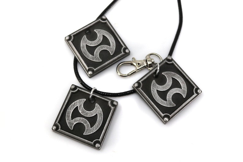 May include: Three silver-colored pendants with a black background and a stylized symbol. The pendants are attached to a black cord necklace.