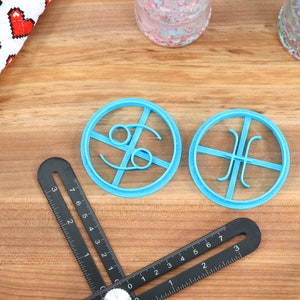 May include: Two blue plastic cookie cutters shaped like zodiac signs. The cutter on the left is shaped like the Cancer zodiac sign and the cutter on the right is shaped like the Pisces zodiac sign.