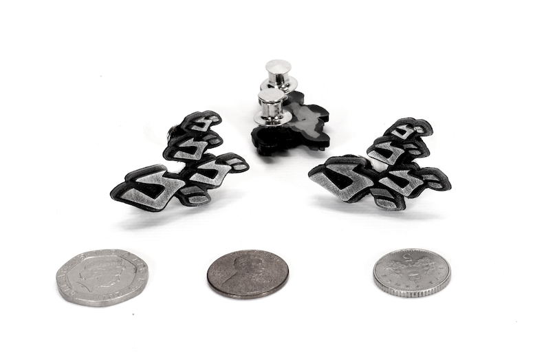 May include: Two silver and black enamel pins with a stylized design. The pins are attached to a white background.