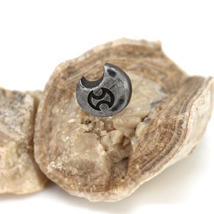 May include: A small, silver-coloured, crescent-shaped pendant with a black symbol in the centre, resting on a piece of light brown rock.