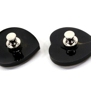 May include: Two black heart-shaped pins with silver metal clasps. The pins are shiny and have a smooth surface.