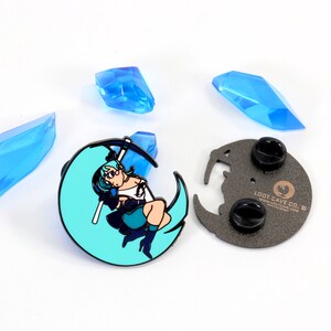 May include: A blue and black enamel pin featuring a cartoon character holding a scythe, standing on a crescent moon. The pin is on a white background with blue crystals. The back of the pin is black with the text "Loot Cave Co."