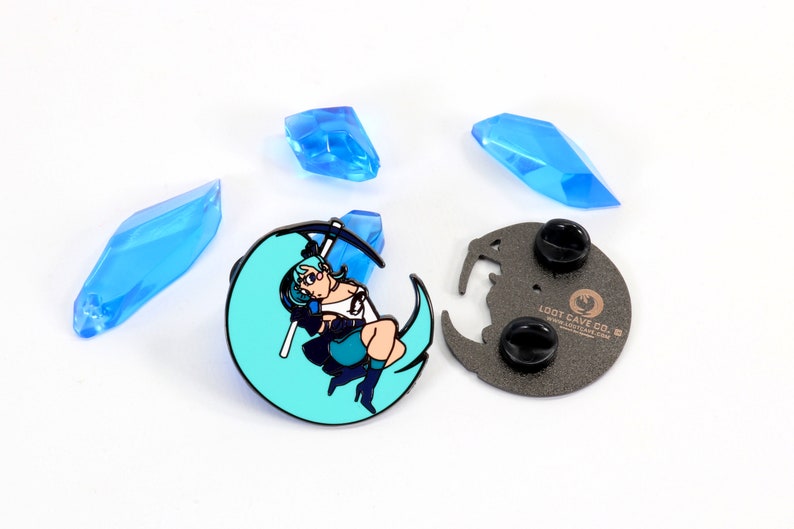 May include: A blue and black enamel pin featuring a cartoon character holding a scythe, standing on a crescent moon. The pin is on a white background with blue crystals. The back of the pin is black with the text "Loot Cave Co."