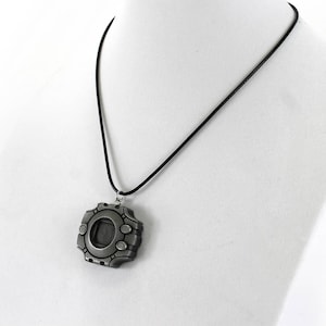 Puede incluir: Un collar con un colgante plateado con un dise&ntilde;o detallado que se asemeja a un dispositivo digital. El colgante tiene una ventana circular y un cord&oacute;n negro.