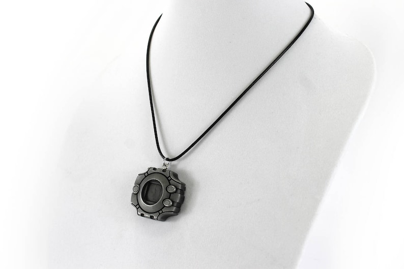 Puede incluir: Un collar con un colgante plateado con un dise&ntilde;o detallado que se asemeja a un dispositivo digital. El colgante tiene una ventana circular y un cord&oacute;n negro.