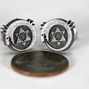 May include: A pair of silver cufflinks featuring a stylized star of David with a serpent wrapped around it. The cufflinks are about the size of a US penny.