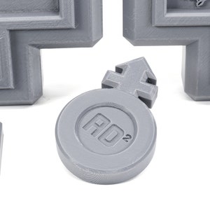 May include: A gray plastic token with a raised arrow symbol and the text "AD2" printed on it.