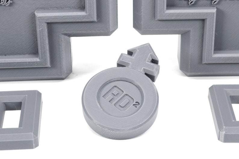 May include: A gray plastic token with a raised arrow symbol and the text "AD2" printed on it.