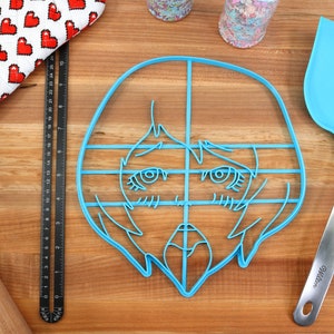 May include: A blue plastic cookie cutter shaped like a cartoon face with large eyes and a grid pattern. The cutter is on a wooden surface.