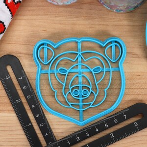 May include: A blue plastic bear-shaped cookie cutter with a grid pattern inside. The cutter is on a wooden surface.