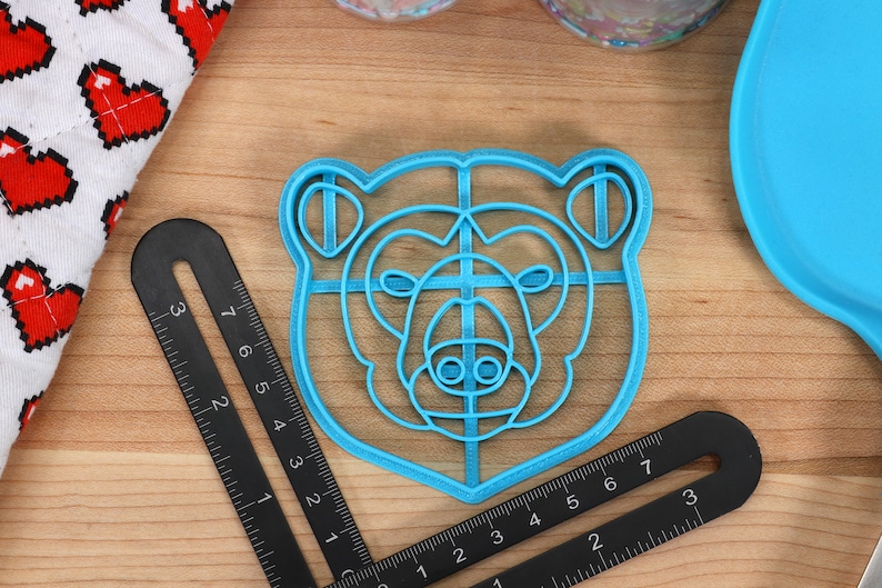 May include: A blue plastic bear-shaped cookie cutter with a grid pattern inside. The cutter is on a wooden surface.