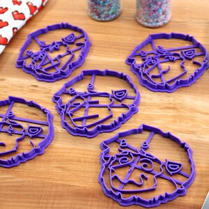 May include: Five purple plastic cookie cutters shaped like a dog's face. The cutters are on a wooden surface.