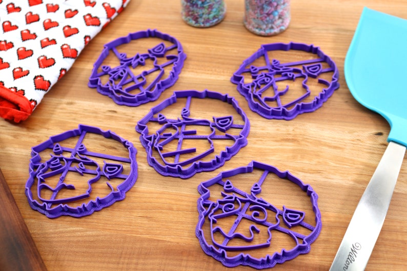 May include: Five purple plastic cookie cutters shaped like a dog's face. The cutters are on a wooden surface.