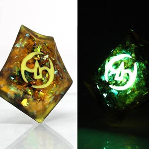 May include: A gold and green iridescent dice with a white symbol of a crescent moon and three stars. The dice glows green in the dark.