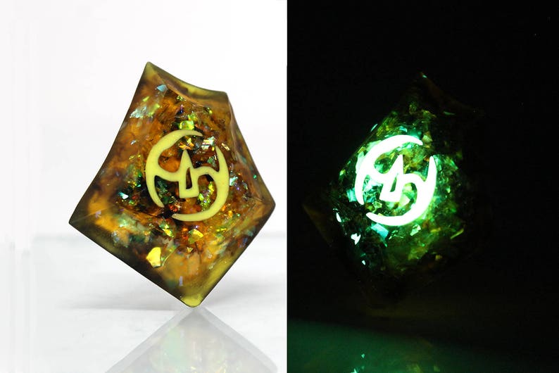 May include: A gold and green iridescent dice with a white symbol of a crescent moon and three stars. The dice glows green in the dark.