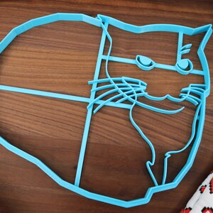 May include: A blue plastic cookie cutter shaped like a cat's face. The cat has a grumpy expression with a raised eyebrow and a slight smirk.