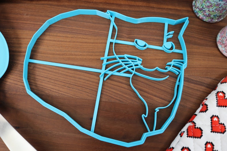 May include: A blue plastic cookie cutter shaped like a cat's face. The cat has a grumpy expression with a raised eyebrow and a slight smirk.