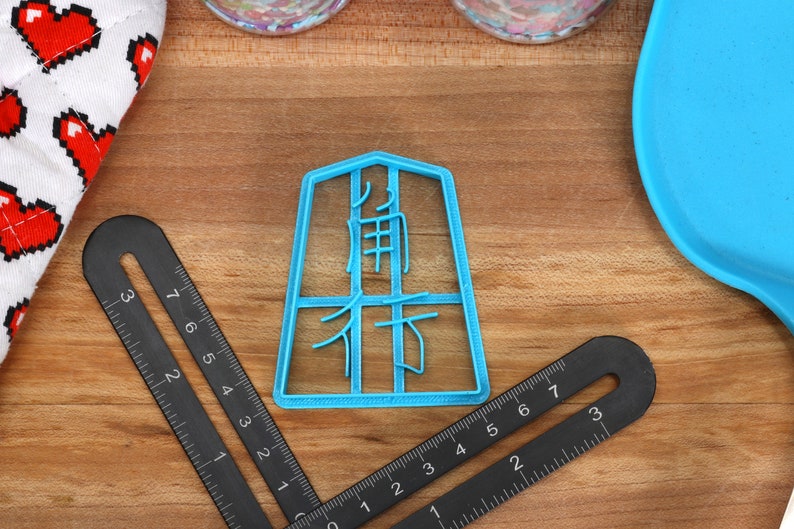 May include: A light blue plastic cookie cutter with Japanese characters is centered on a wooden surface. Two black angle rulers with measurement markings are positioned around the cutter. A white cloth with red pixelated hearts is visible.
