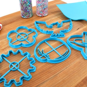 May include: A collection of blue plastic cookie cutters in various shapes, including a flower, gear, and bird, are arranged on a wooden surface. A blue spatula and a heart-patterned oven mitt are also present, along with jars of sprinkles.