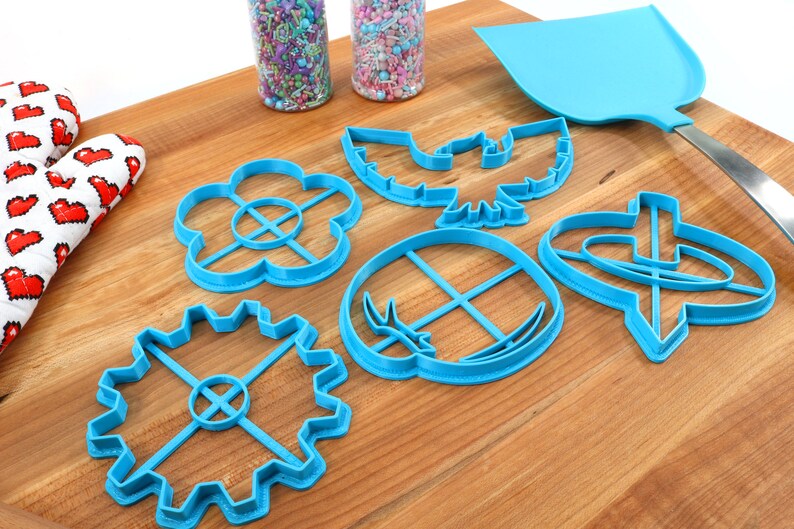 May include: A collection of blue plastic cookie cutters in various shapes, including a flower, gear, and bird, are arranged on a wooden surface. A blue spatula and a heart-patterned oven mitt are also present, along with jars of sprinkles.