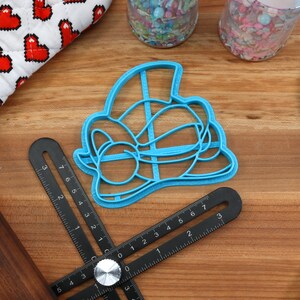 May include: A blue plastic cookie cutter shaped like a cartoon character with a hat and bow tie. The cutter is on a wooden surface with a ruler and rolling pin.