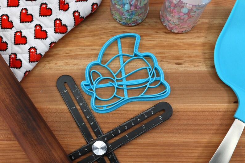 May include: A blue plastic cookie cutter shaped like a cartoon character with a hat and bow tie. The cutter is on a wooden surface with a ruler and rolling pin.