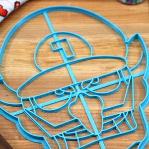 May include: A blue plastic cookie cutter shaped like a cartoon character with a large head, a single eye, and a wide mouth. The cutter is on a wooden surface.