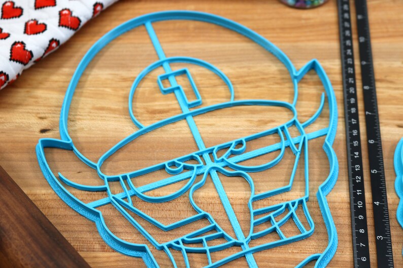 May include: A blue plastic cookie cutter shaped like a cartoon character with a large head, a single eye, and a wide mouth. The cutter is on a wooden surface.
