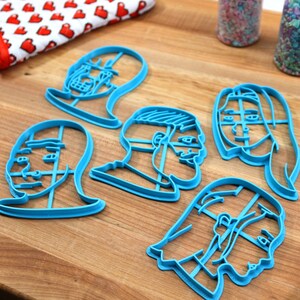 Op de afbeelding: Zes blauwe plastic koekjesvormen in de vorm van verschillende gezichten. De vormen liggen op een houten oppervlak.