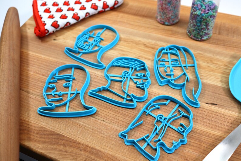Op de afbeelding: Zes blauwe plastic koekjesvormen in de vorm van verschillende gezichten. De vormen liggen op een houten oppervlak.