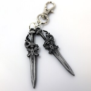 May include: A silver-colored keychain featuring two decorative sword charms. Each sword has an ornate handle and blade, connected to a silver clasp. The charms are detailed with decorative elements.