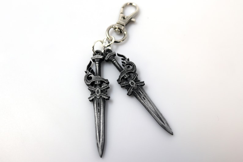 May include: A silver-colored keychain featuring two decorative sword charms. Each sword has an ornate handle and blade, connected to a silver clasp. The charms are detailed with decorative elements.