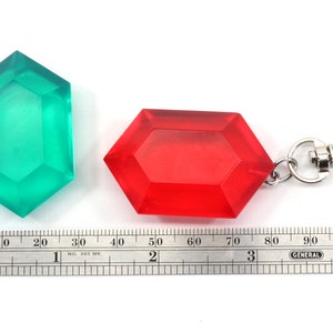 May include: Two plastic Triforce keychains, one green and one red. The red keychain has a silver metal clasp. A ruler is in the bottom of the image to show the size of the keychains.
